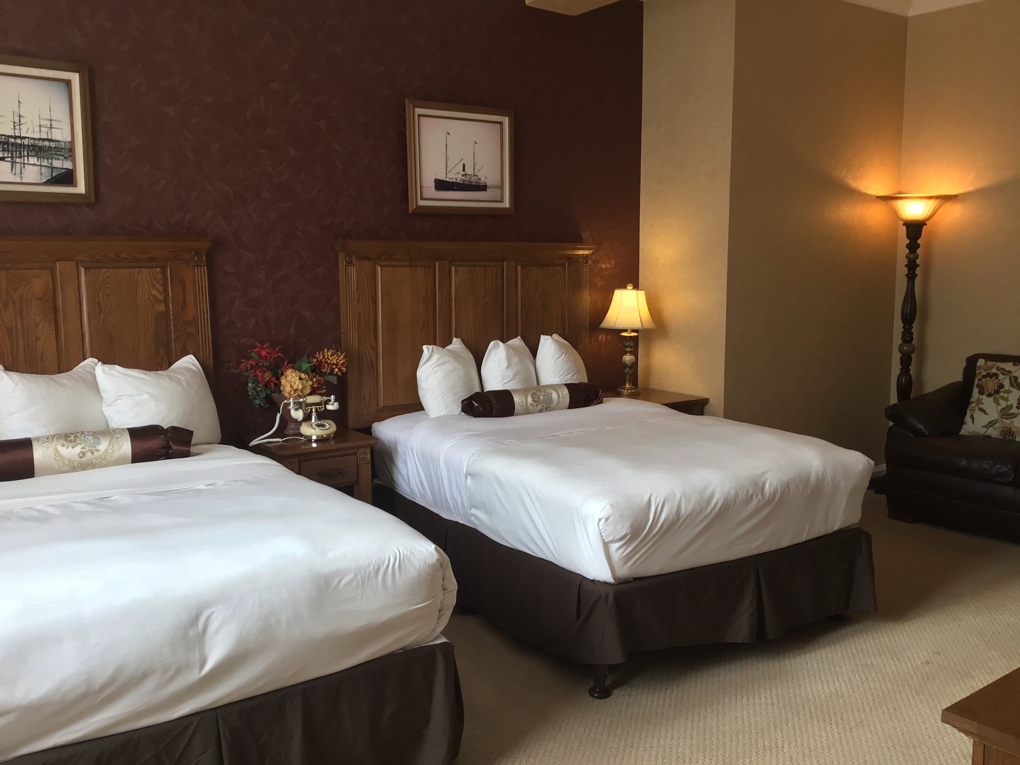 A hotel room with two beds, wooden headboards, a nightstand with a lamp, and a floral armchair.