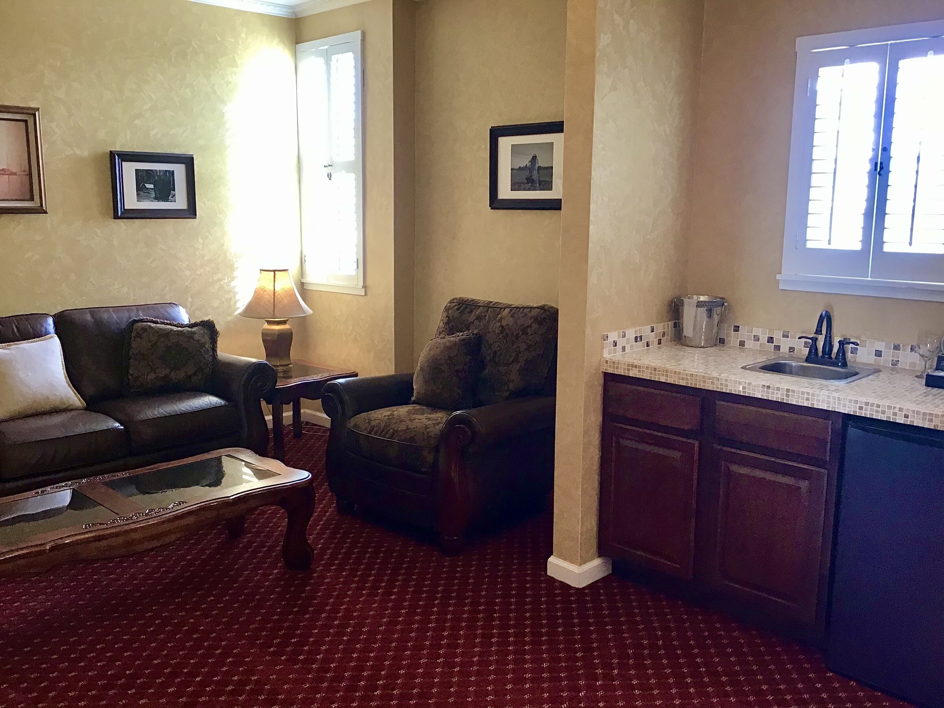 A living room with a sofa, armchair, and coffee table. There is a window with blinds, a lamp, and a kitchen area with a sink and cabinets.