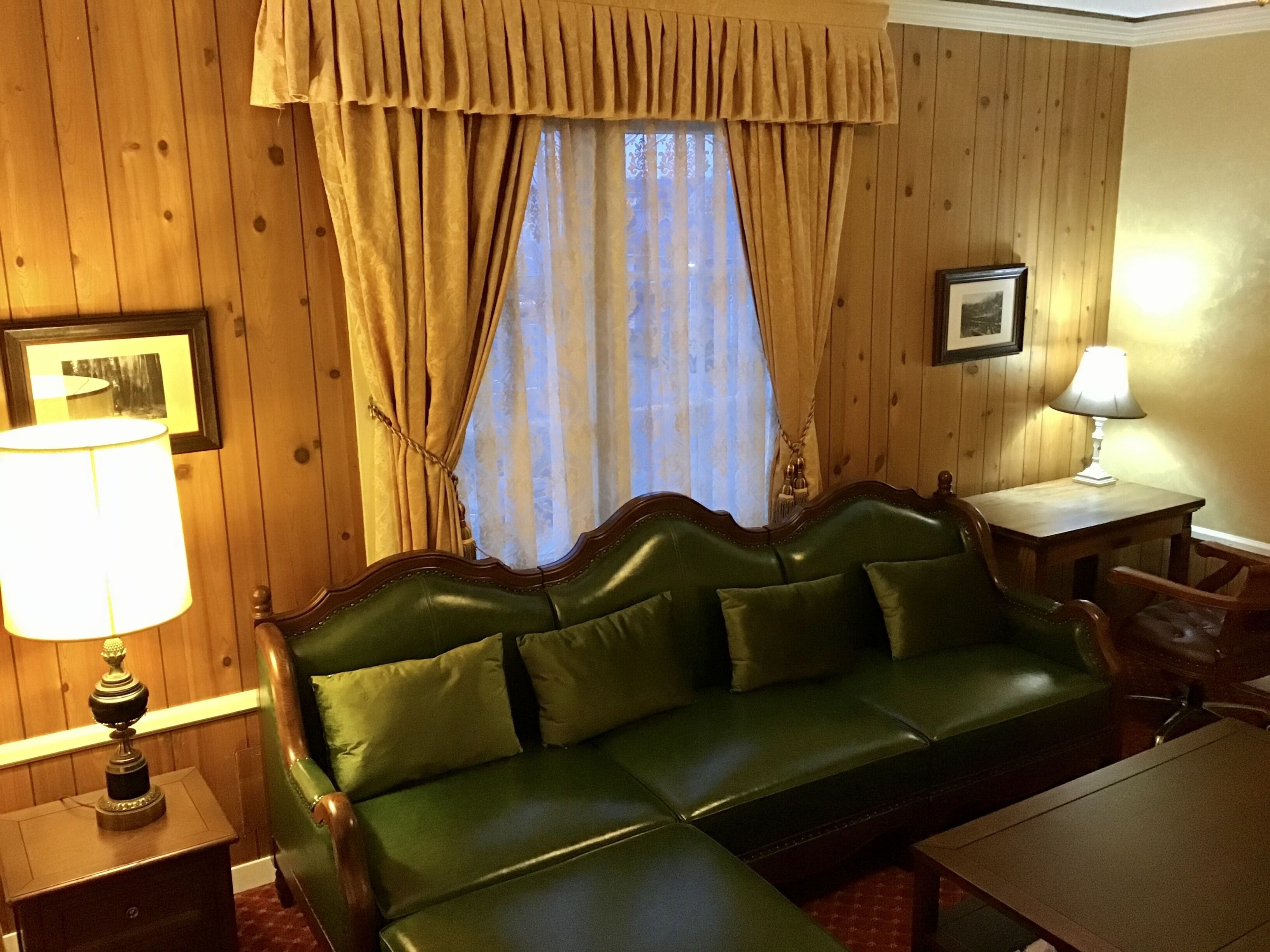 A room with a green leather sofa, a wooden table with a lamp, and a window with curtains.