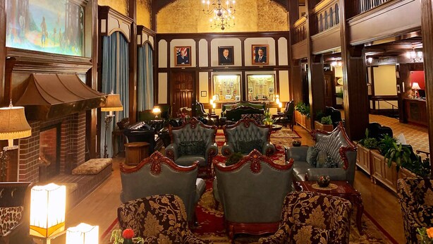 A grand lobby with a chandelier, ornate chairs, a fireplace, and framed portraits on the walls.