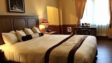 Habitación de hotel con una cama grande, un cabecero de madera, una mesita de noche con lámpara, un escritorio con silla y una ventana con cortinas.