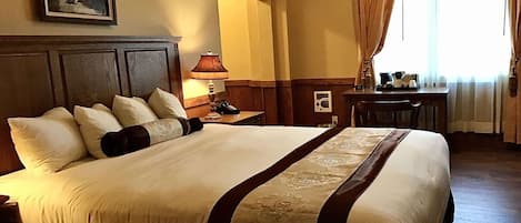 Habitación de hotel con una cama grande, un cabecero de madera, una mesita de noche con lámpara, un escritorio con silla y una ventana con cortinas.
