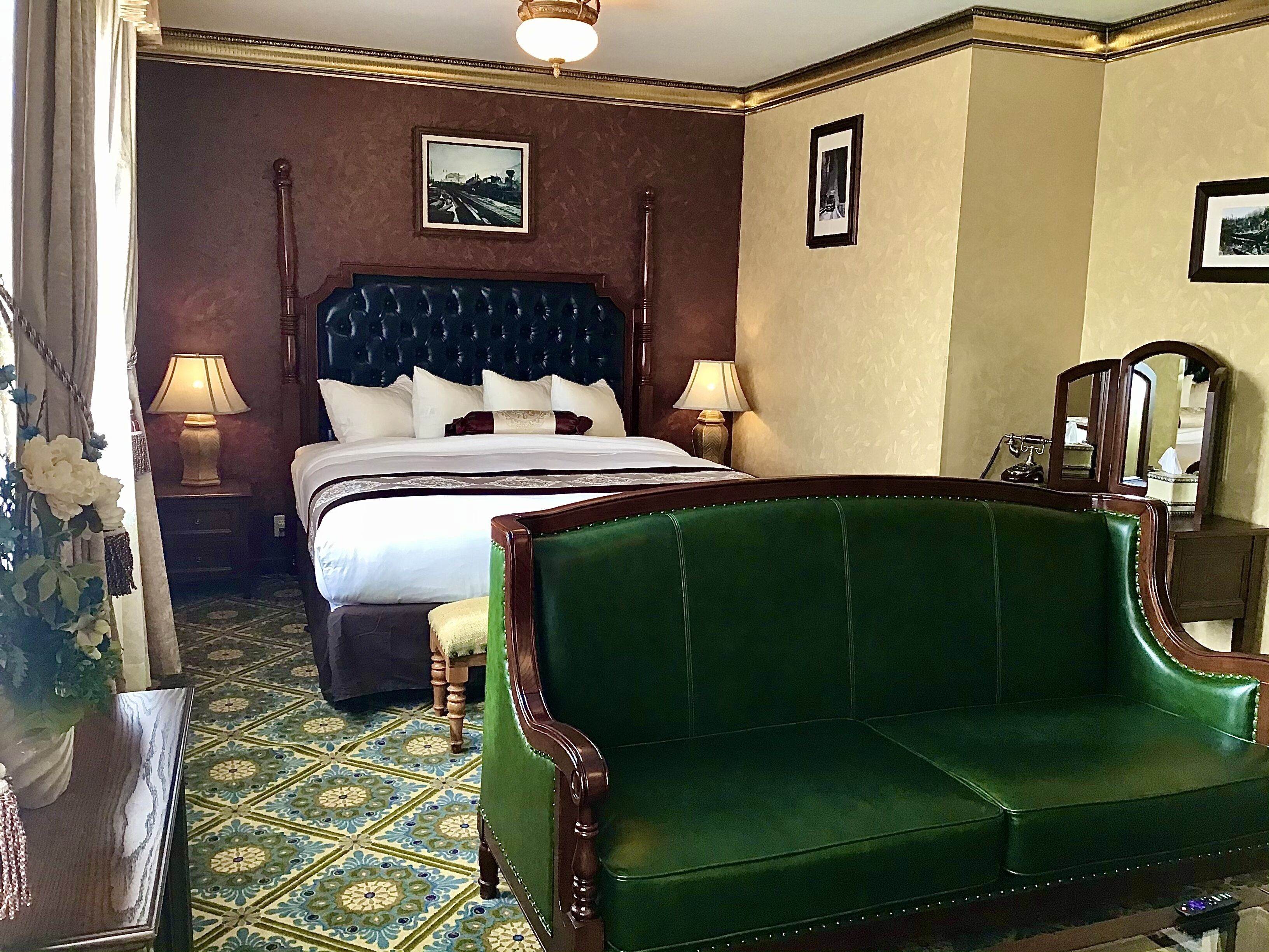 A hotel room with a large bed, two bedside lamps, a green sofa, a patterned floor, and a mirror.