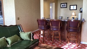 A room with a green sofa, a wooden coffee table, and a patterned red carpet. There are four wooden chairs with cushioned seats. A small bar area with a lamp and a telephone is visible.