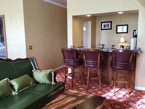 A room with a green sofa, a wooden coffee table, and a patterned red carpet. There are four wooden chairs with cushioned seats. A small bar area with a lamp and a telephone is visible.