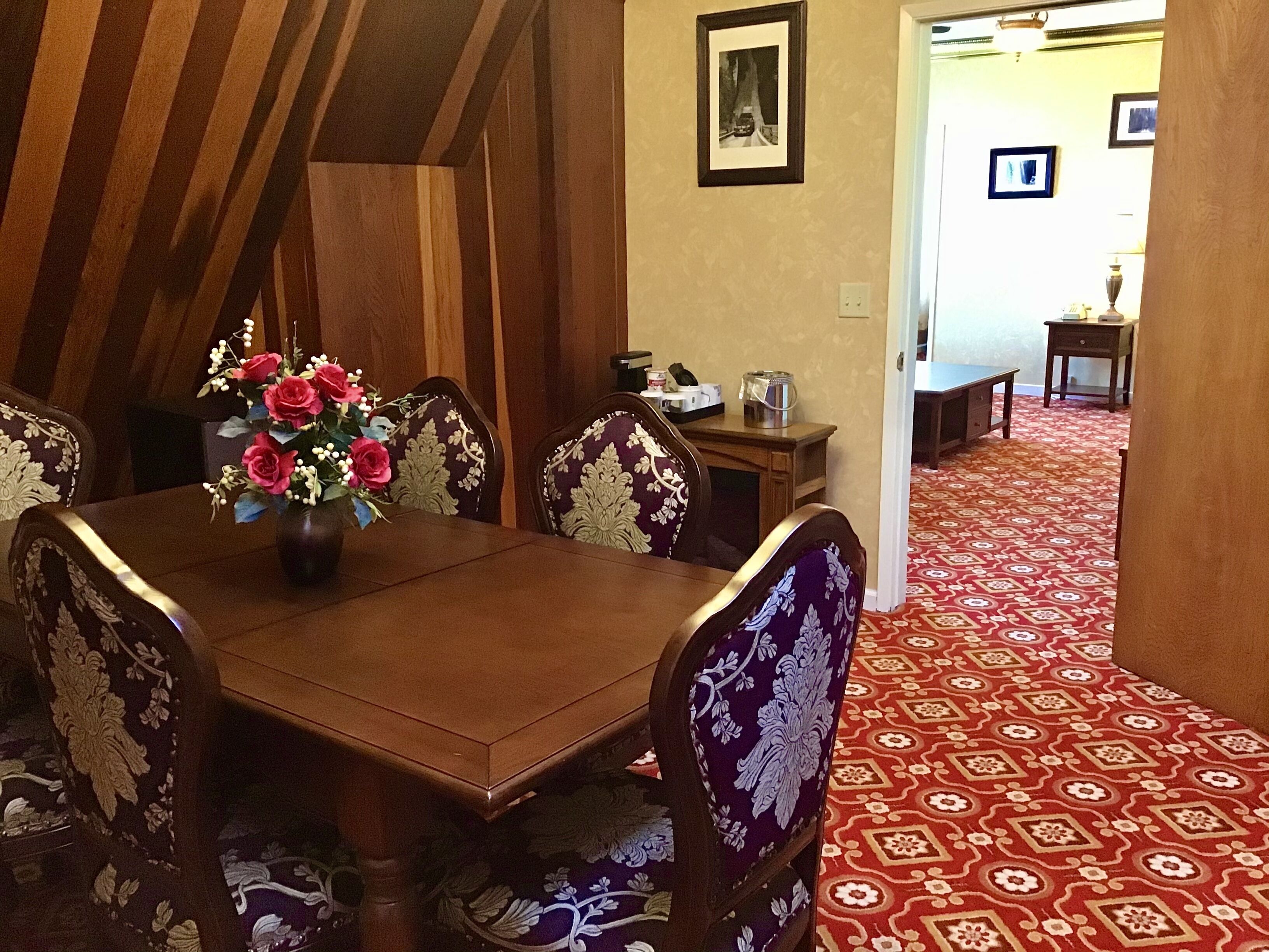 A dining area with a wooden table and chairs, a vase of flowers, and a patterned carpet.