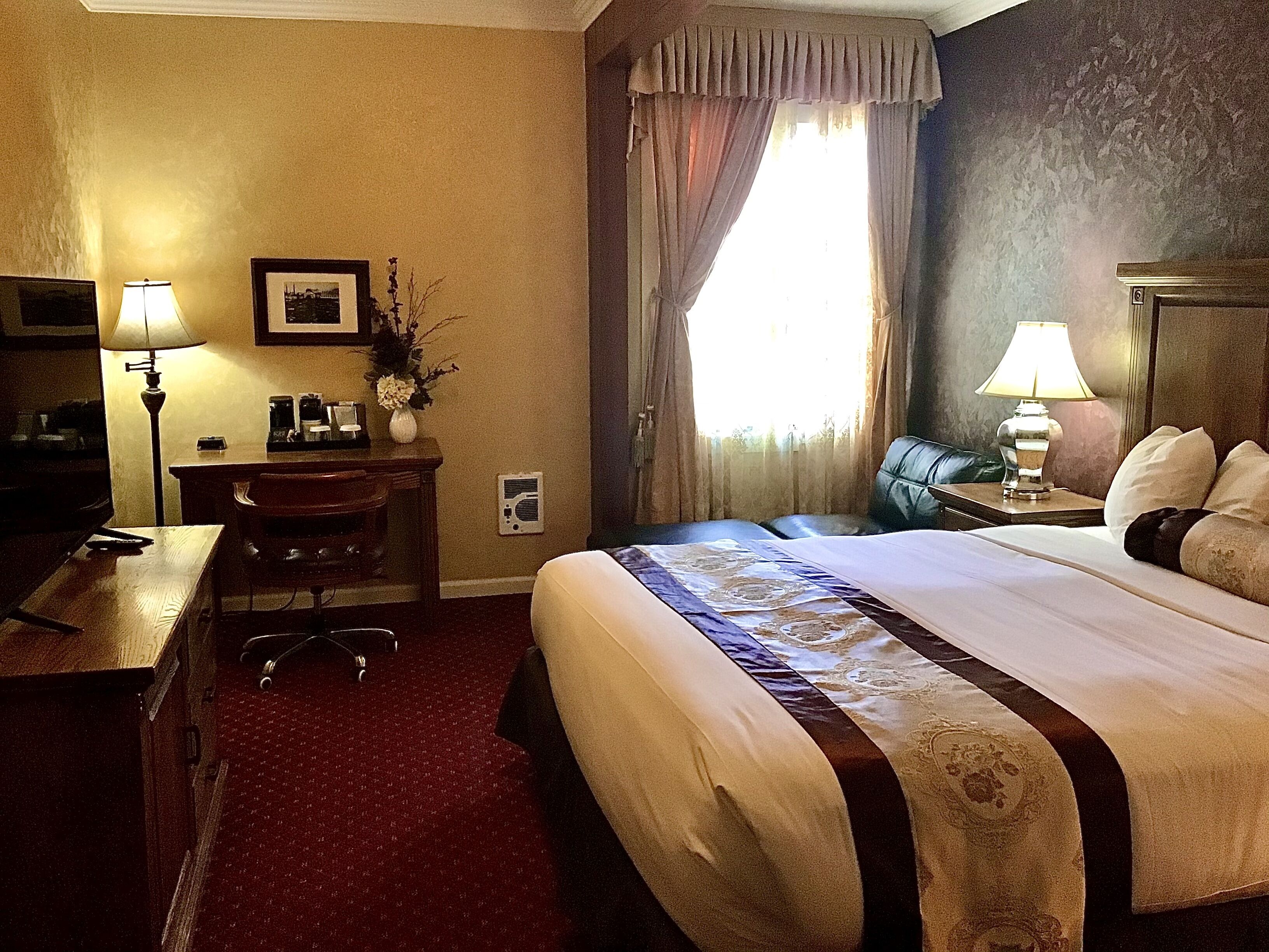 Habitación de hotel con una cama grande, un escritorio, una silla, una mesita de noche, una lámpara, un cuadro en la pared y una ventana con cortinas.