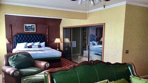 A hotel room with a large bed, a green armchair, a green sofa, and a glass door leading to a balcony.