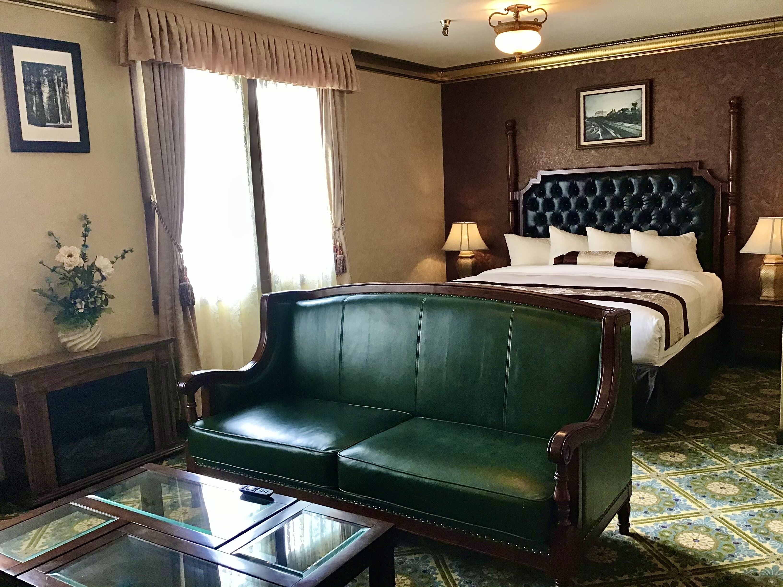 Quarto de hotel com sofá de couro verde, mesa de centro de vidro, cama com cabeceira escura e mesa de cabeceira com abajur.