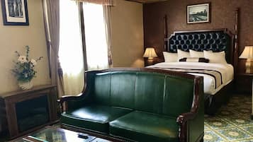 Quarto de hotel com sofá de couro verde, mesa de centro de vidro, cama com cabeceira escura e mesa de cabeceira com abajur.