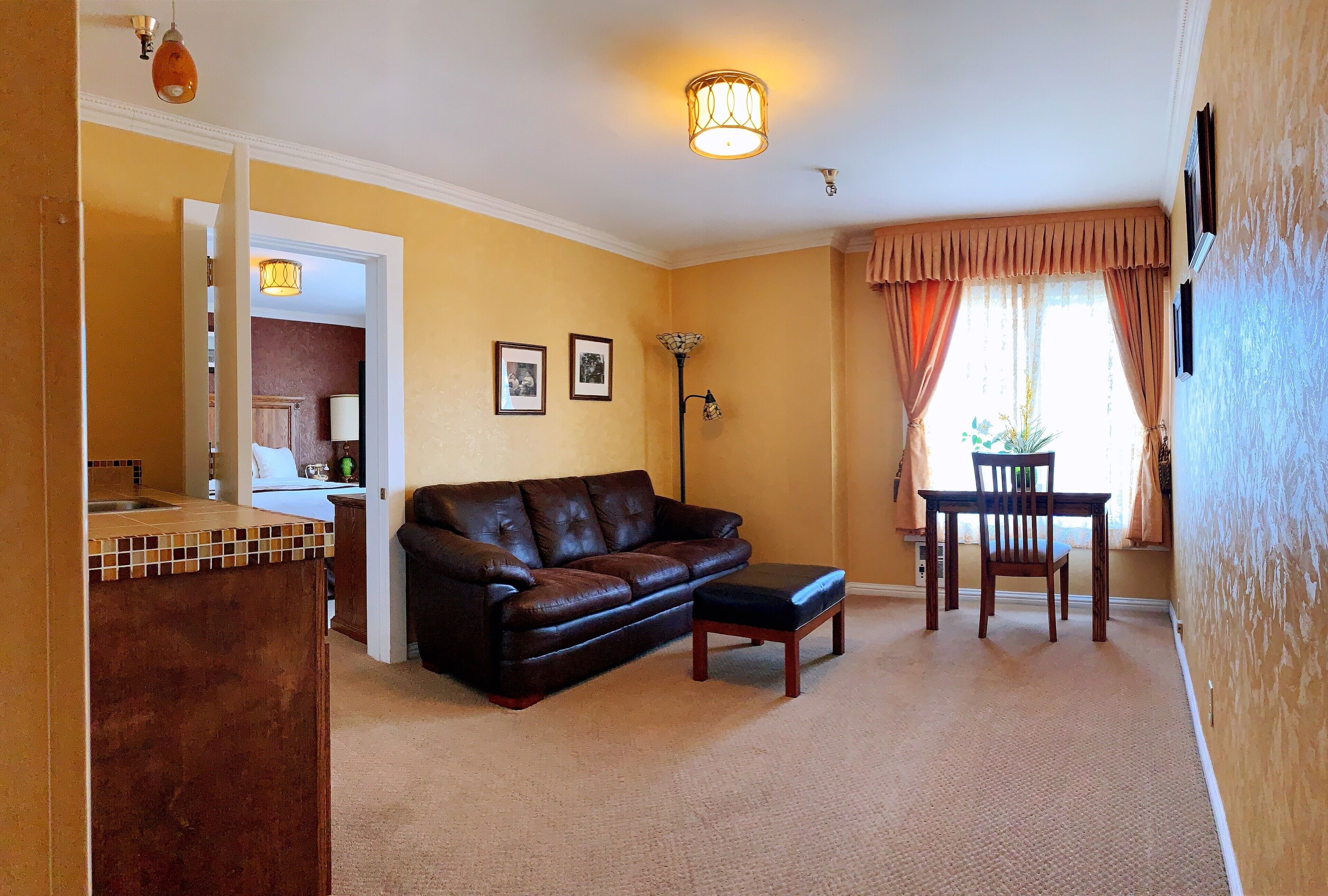 A living room with a brown leather sofa, a matching ottoman, a dining table with chairs, a floor lamp, and a kitchen area visible through an open doorway.