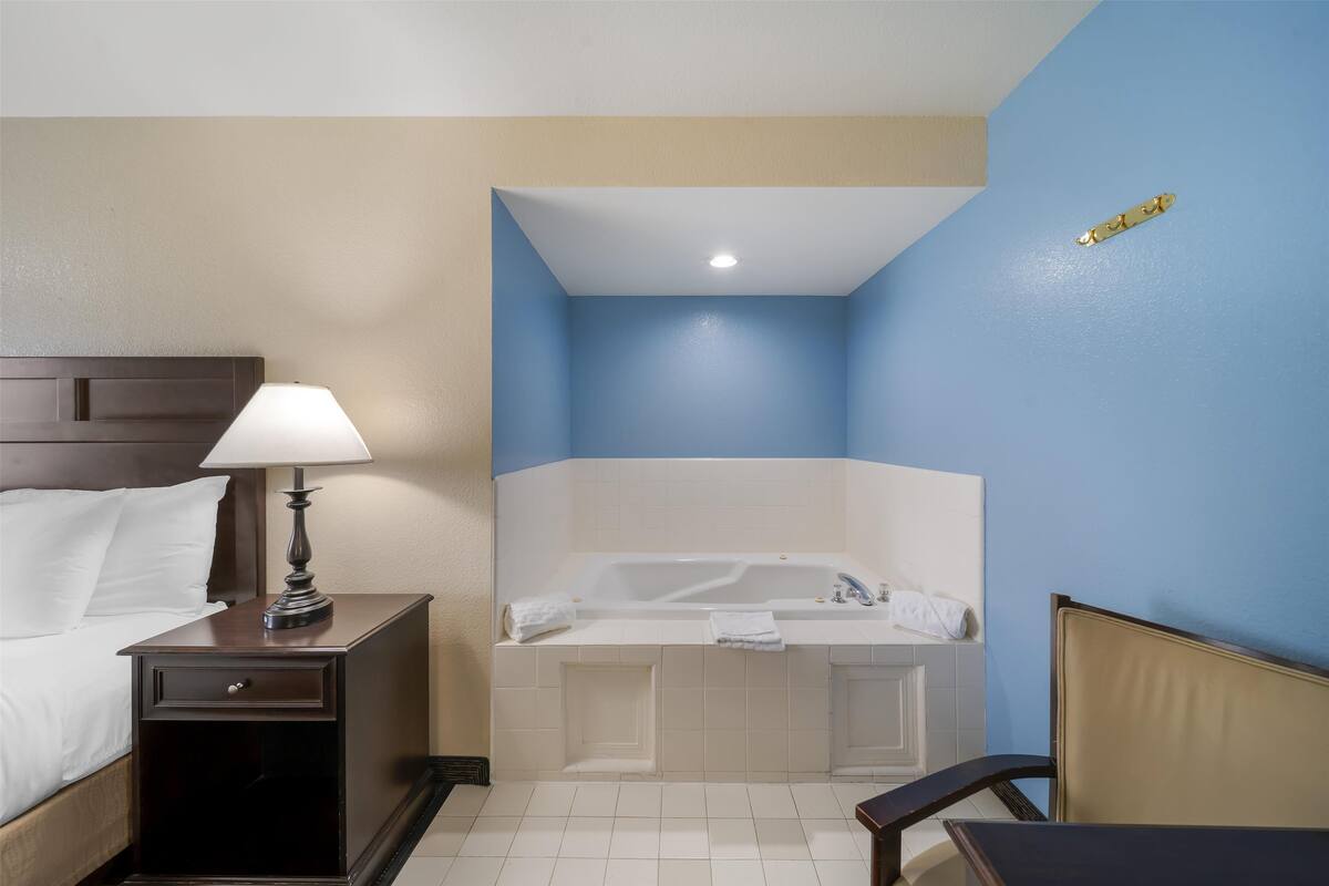 A hotel room with a bed, nightstand, lamp, and a corner bathtub.