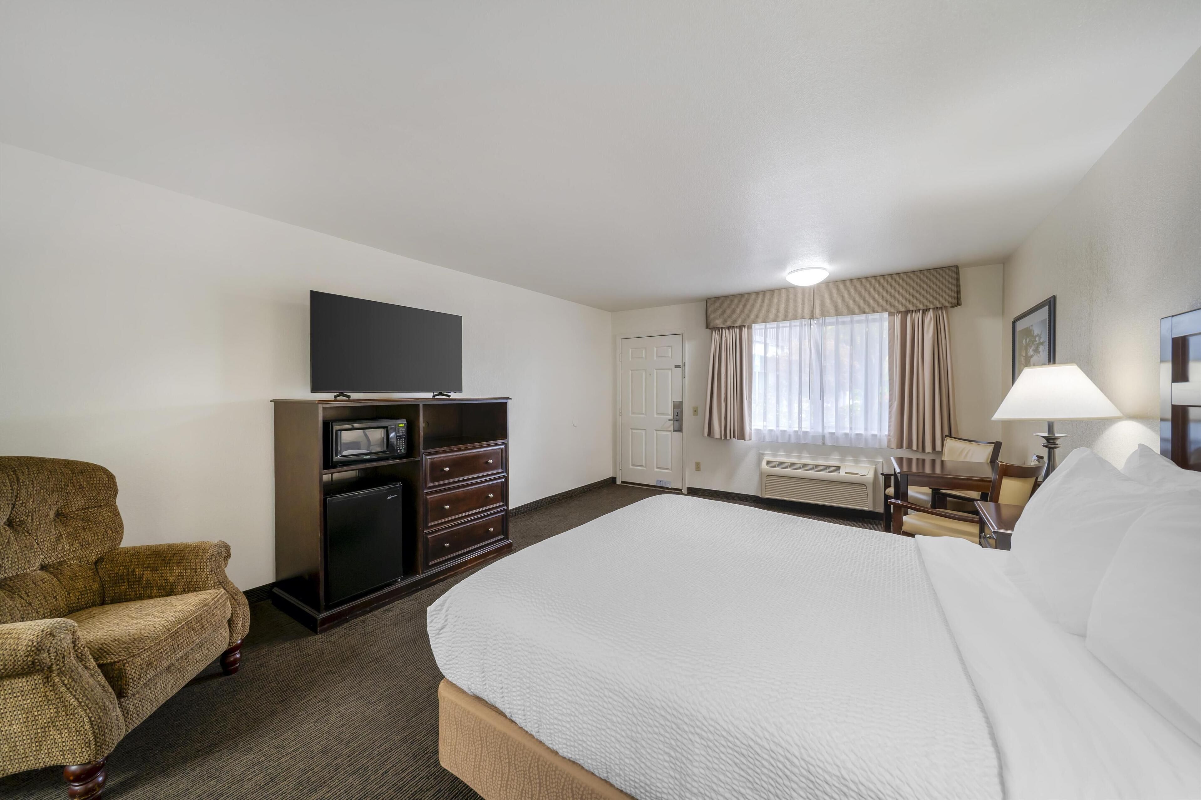 A hotel room with a large bed, a television on a dresser, a chair, a desk with a lamp, and a window with curtains.