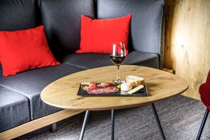 Een ronde houten tafel met een zwarte leistenen serveerplank vol met verschillende kazen, vleeswaren en olijven, vergezeld van een glas rode wijn, geplaatst op een grijze bank met rode kussens.