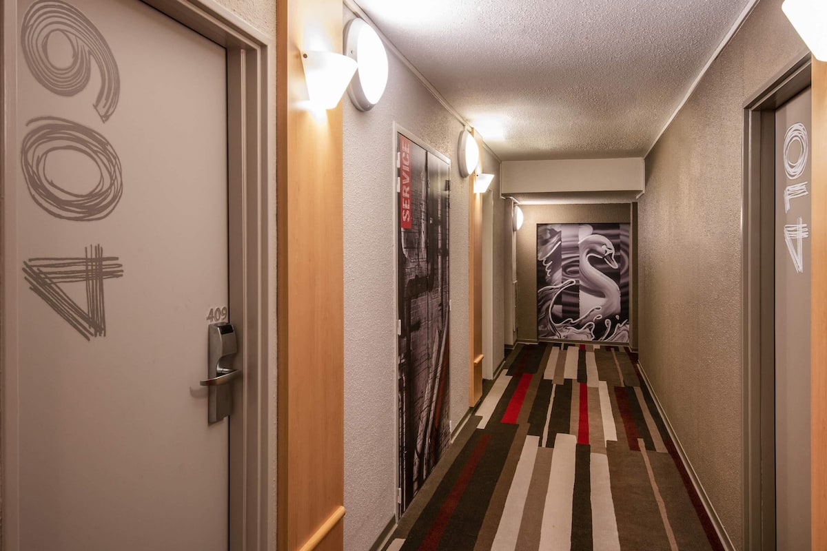 A hallway with striped carpet, wall-mounted light fixtures, and a large abstract painting on the right.