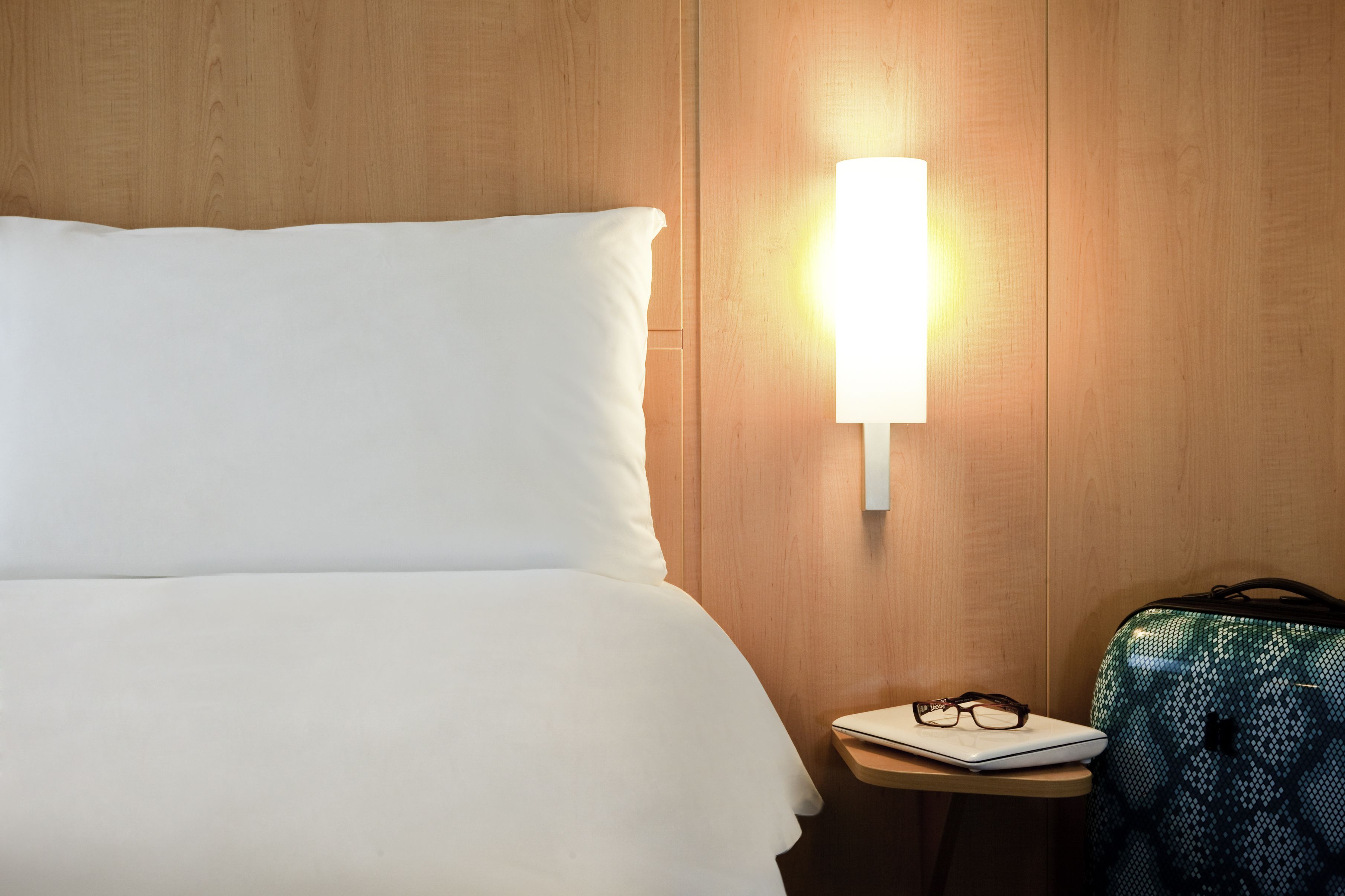 A neatly made bed with white linens, a bedside table with a laptop and glasses, and a suitcase on the floor.