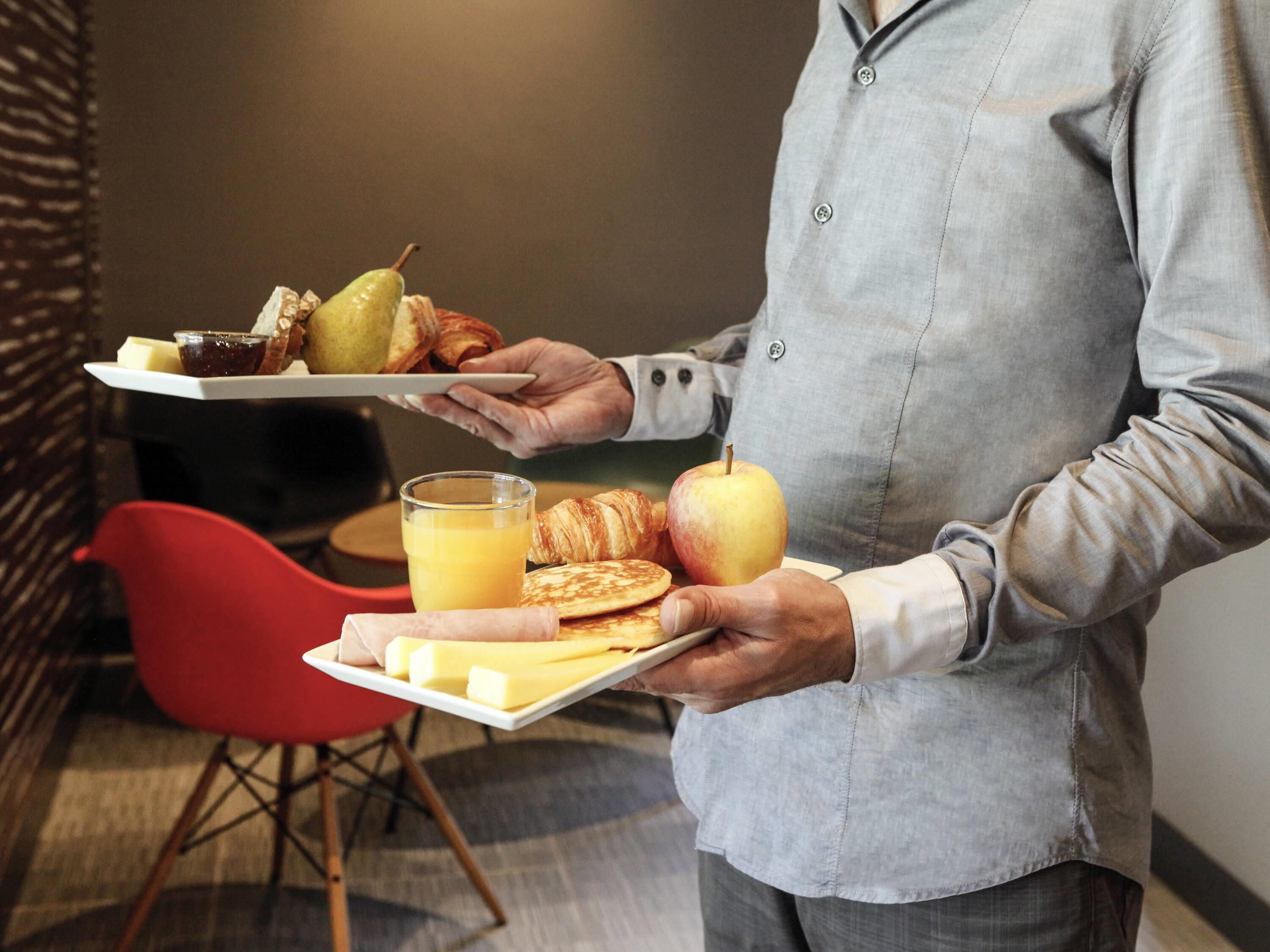 A person holding a tray with breakfast items including toast, a pear, a croissant, an apple, and a glass of orange juice.
