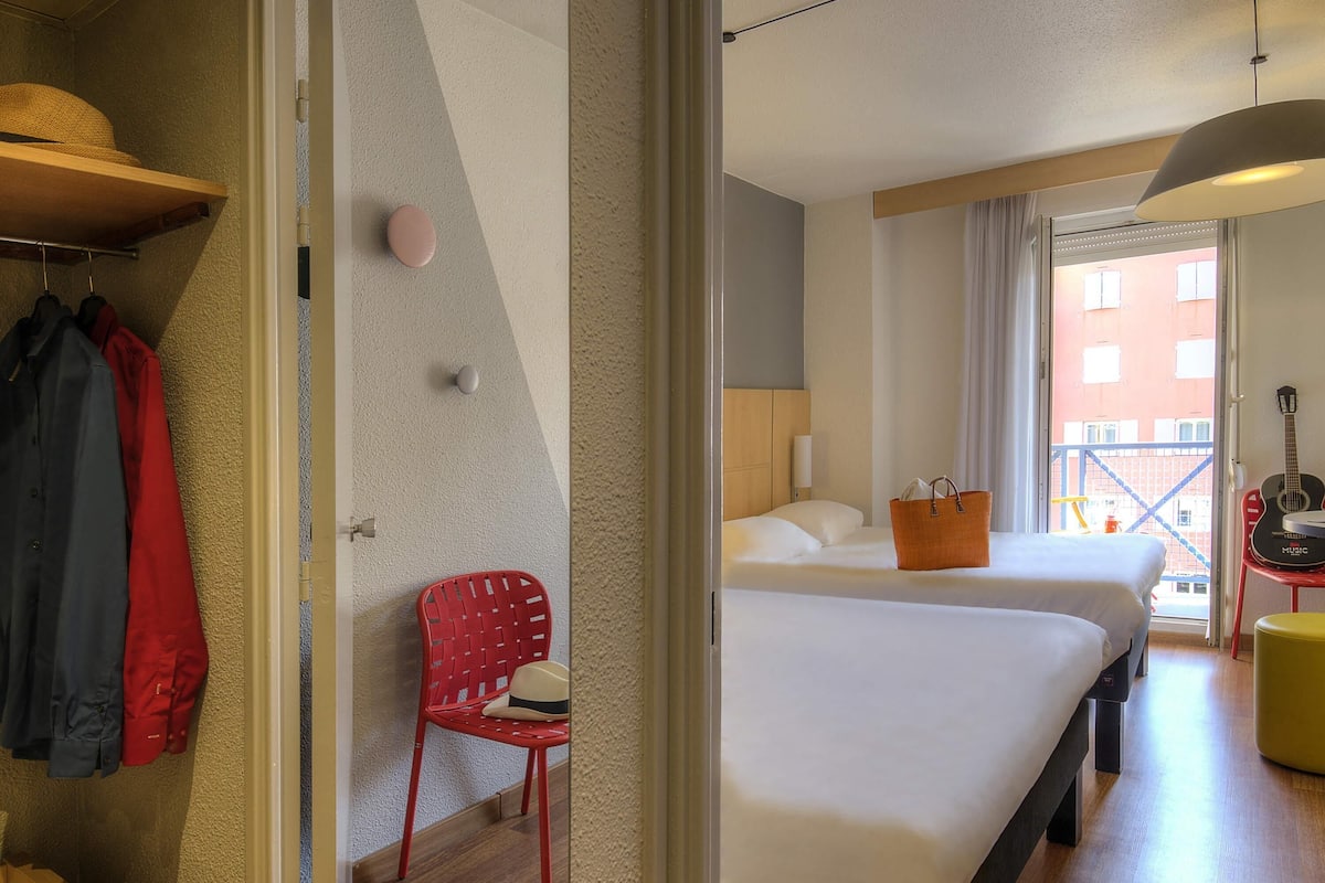 A hotel room with two beds, a red chair, a hanging hat, and a guitar.