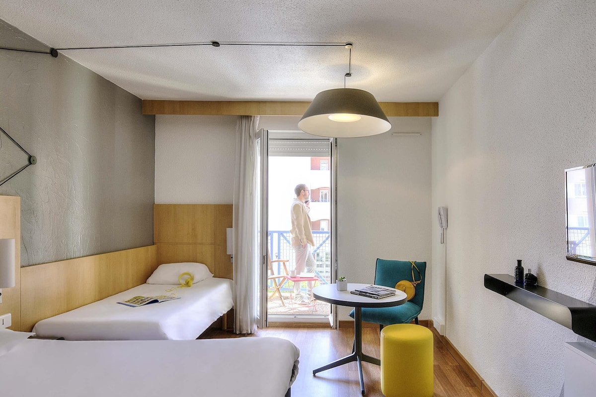 A hotel room with two beds, a small table, a chair, a yellow ottoman, a ceiling light, and a balcony visible through the door.