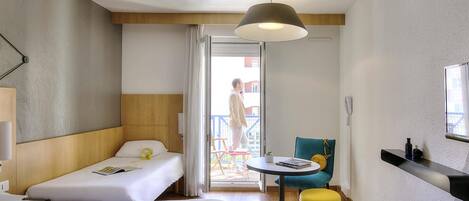 A hotel room with two beds, a small table, a chair, a yellow ottoman, a ceiling light, and a balcony visible through the door.