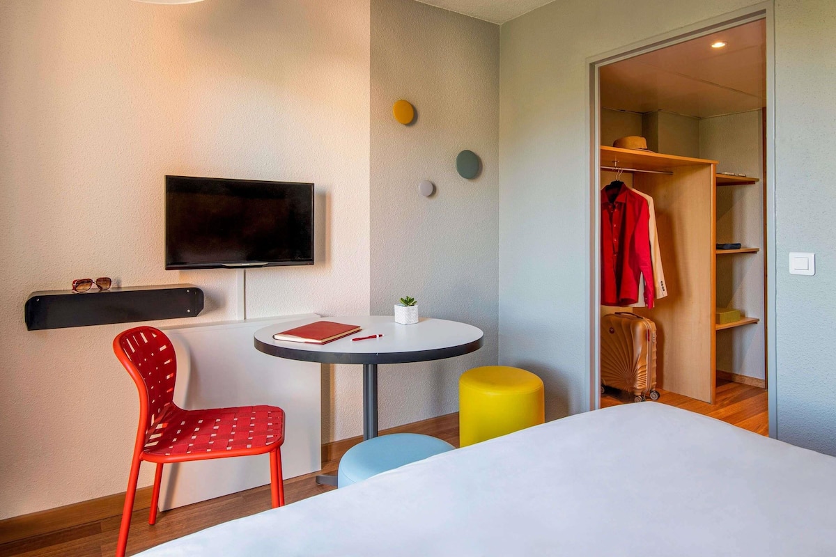 A hotel room with a bed, a TV, a desk with a chair, a small table, and a closet with a red coat.