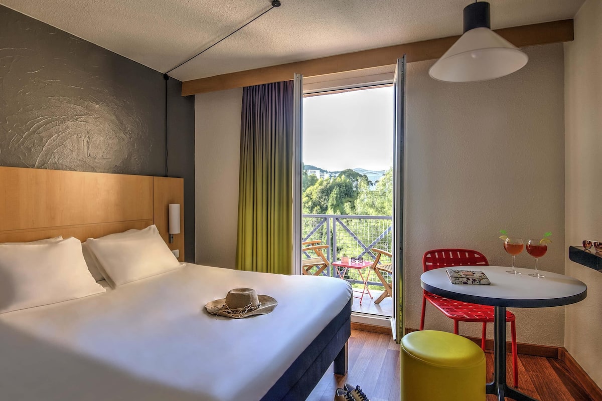 A hotel room with a bed, a small balcony with a table and chairs, and a view of the outdoors.