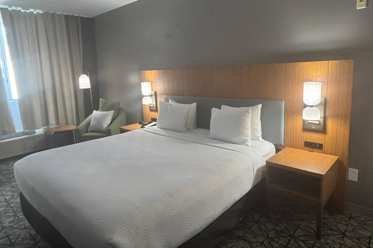 A hotel room with a large bed, two bedside tables, a wall-mounted lamp, and a window with curtains.