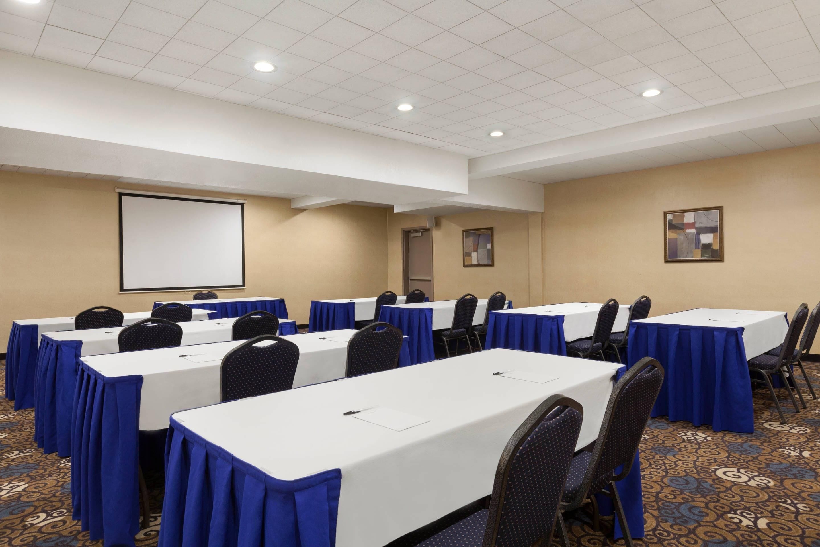 Une salle de conférence équipée de tables, de chaises, d’un écran de projection et d’un sol recouvert de moquette.