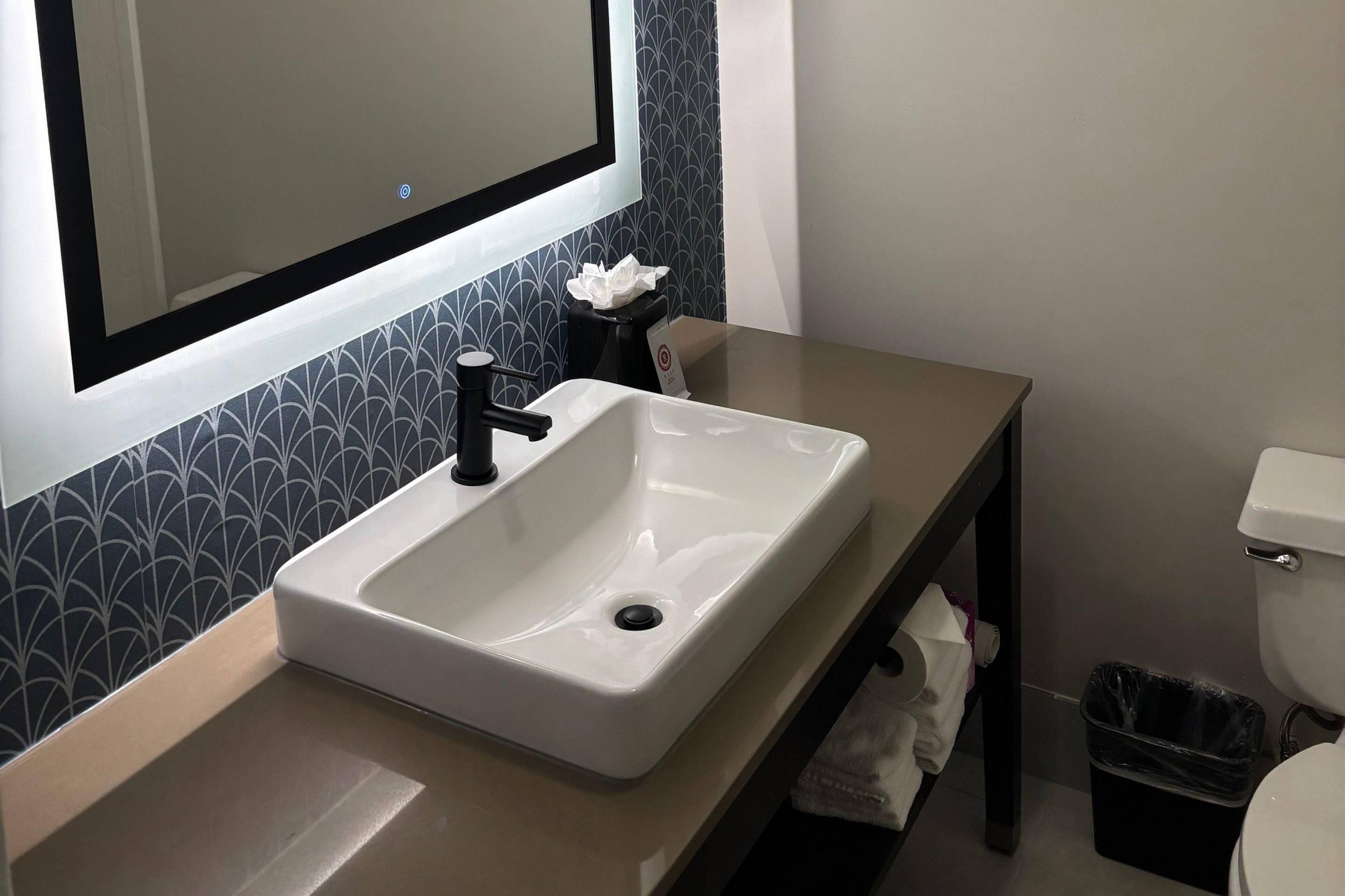 Une salle de bain avec un lavabo blanc, un robinet noir et un miroir au-dessus. On trouve une poubelle noire à côté des toilettes.