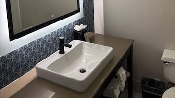 Une salle de bain avec un lavabo blanc, un robinet noir et un miroir au-dessus. On trouve une poubelle noire à côté des toilettes.