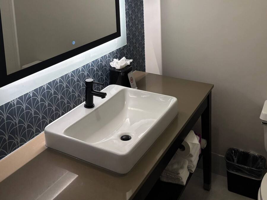 Une salle de bain avec un lavabo blanc, un robinet noir et un miroir au-dessus. On trouve une poubelle noire à côté des toilettes.