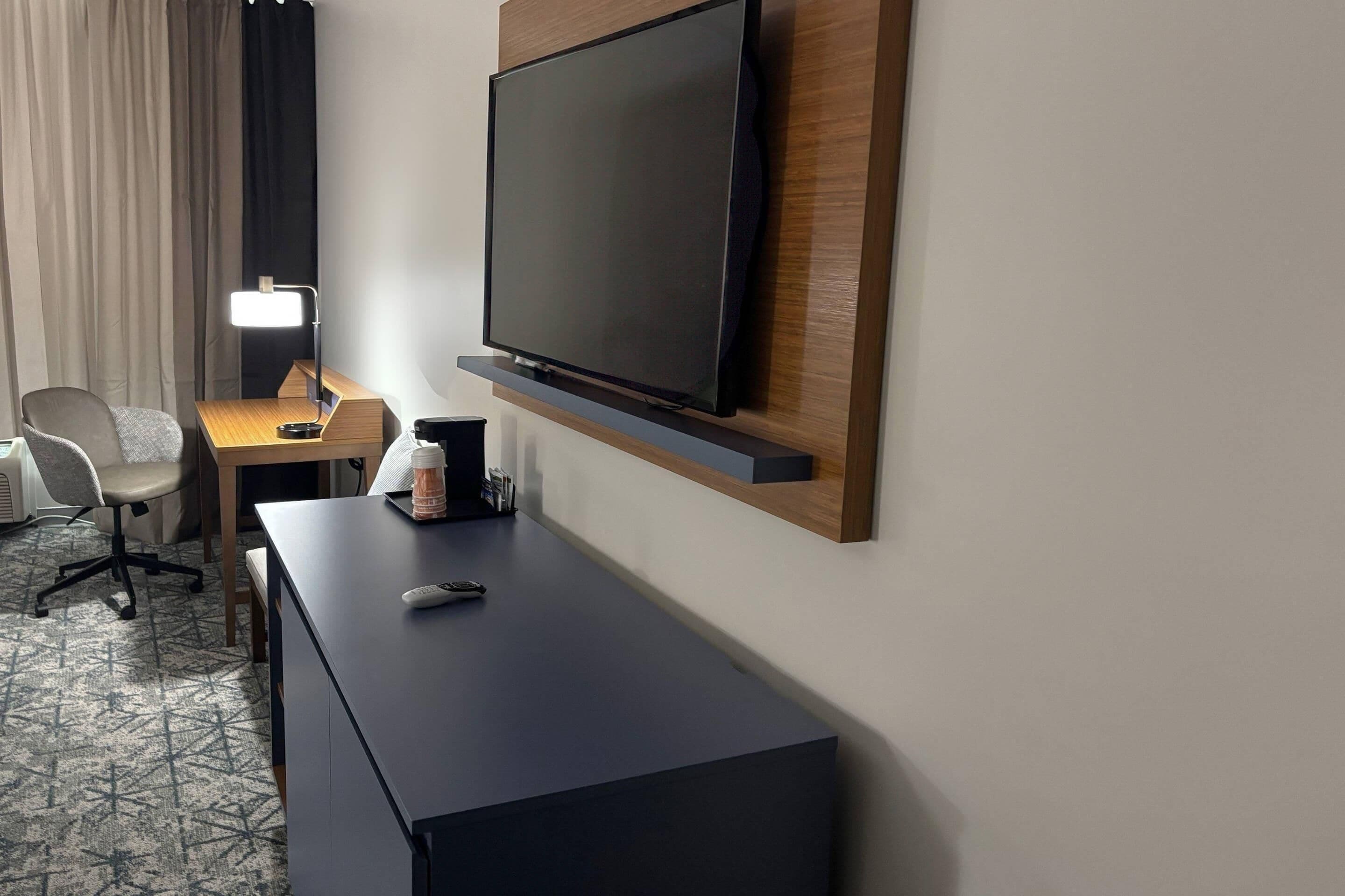 Habitación de hotel con televisión de pantalla plana montada en un panel de madera, un escritorio con lámpara, una silla y una cafetera.