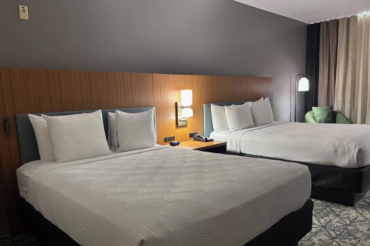A hotel room with two beds, a wooden headboard, a nightstand with a phone, and a lamp.