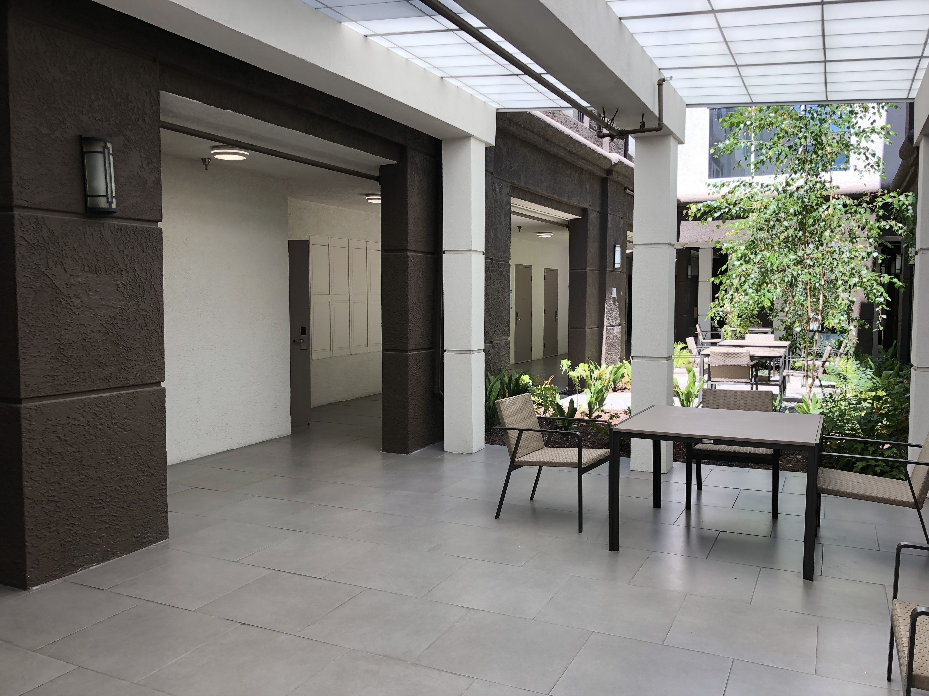 Un espacio interior moderno con techo de vidrio, una zona de estar con mesas y sillas, y vistas a un jardín con plantas y un árbol.