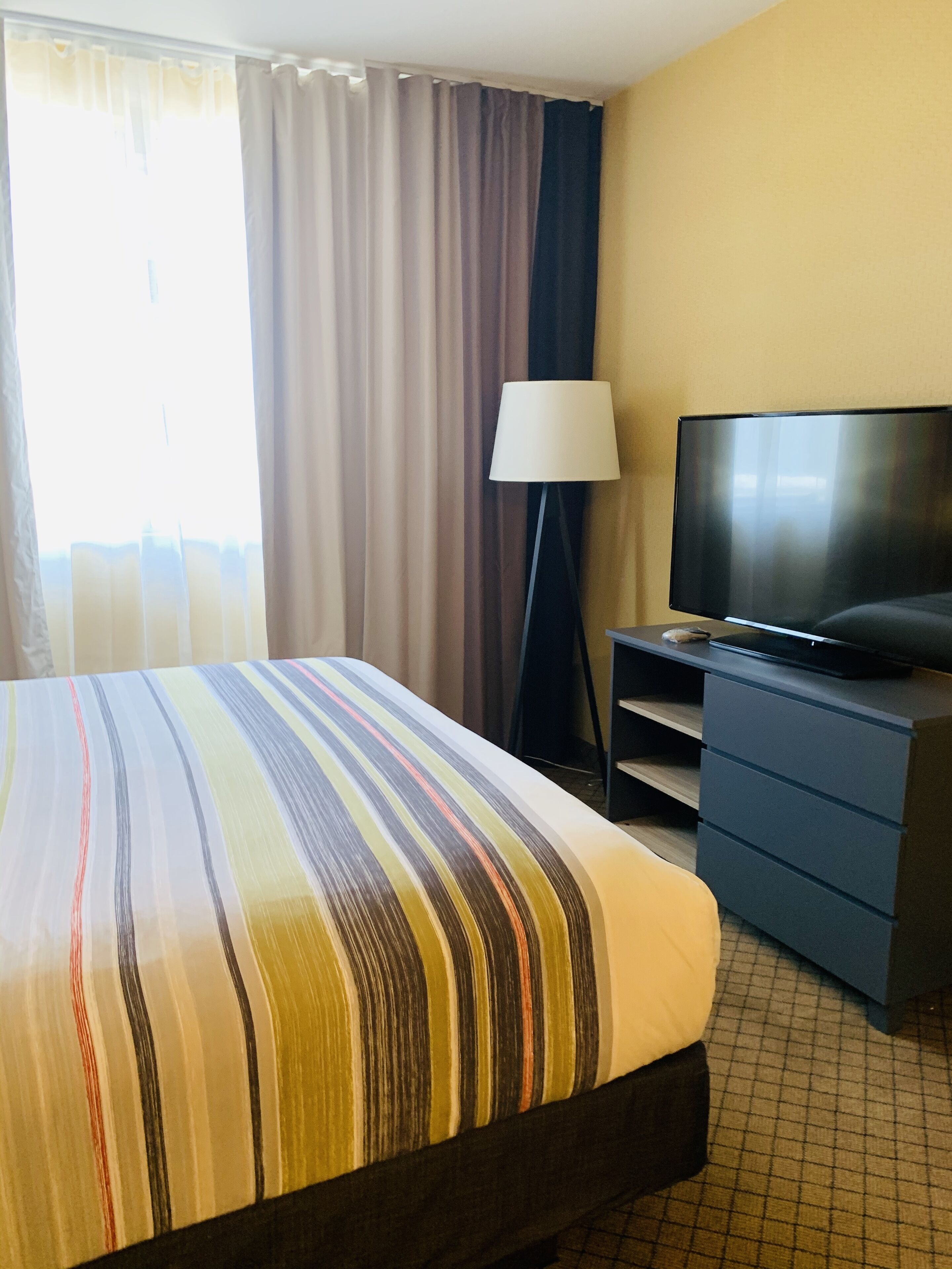 Quarto de hotel com uma cama, televisão em um suporte, escrivaninha e luminária.
