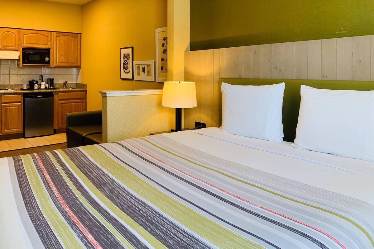 A hotel room with a bed, a nightstand with a lamp, a small desk, a microwave, and a refrigerator in the kitchen area.