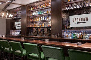 Zona de bar con varias estanterías llenas de botellas de licores, un letrero que dice 'JACKDAW' y taburetes de bar tapizados en verde.