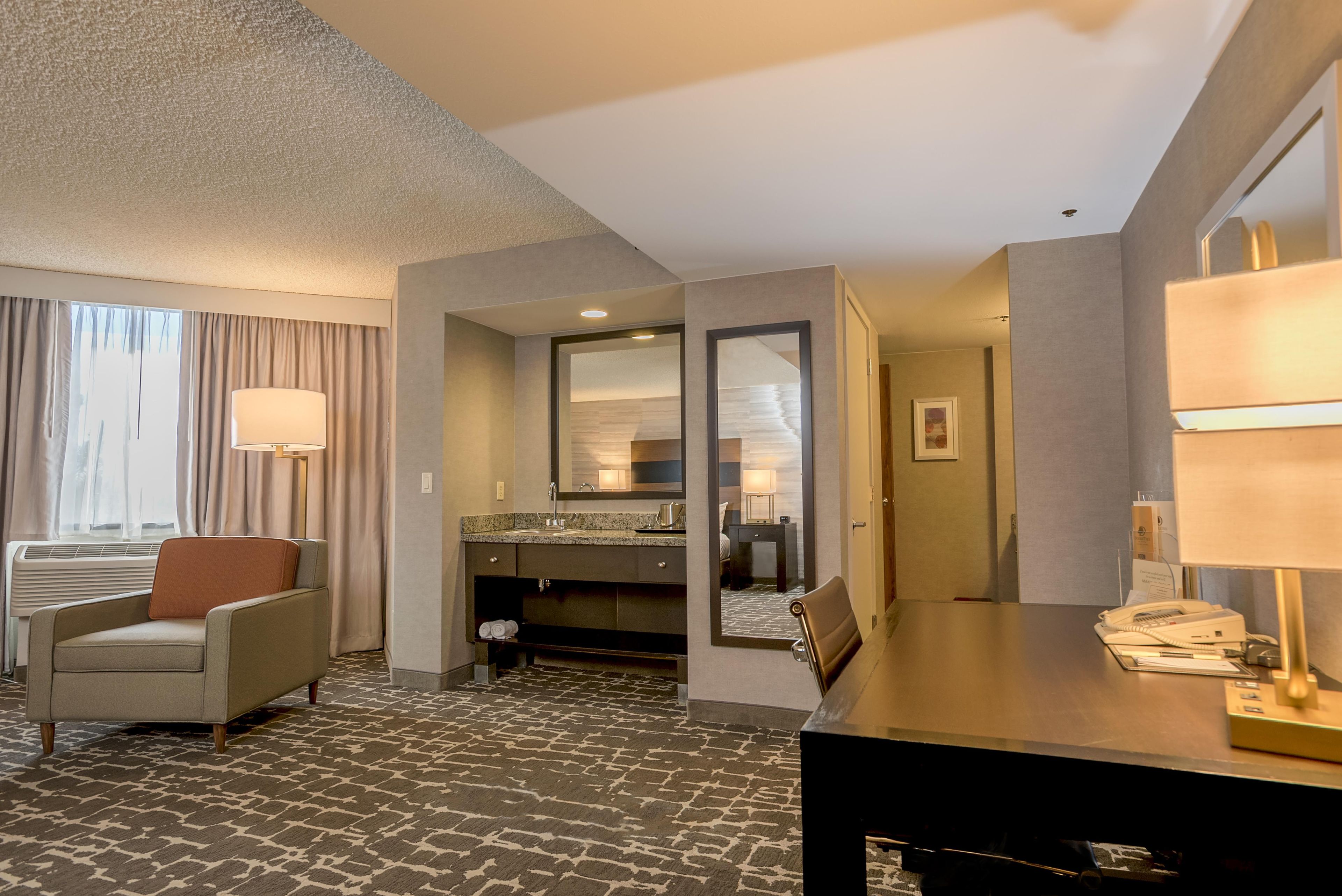A hotel room with a bed, a desk, a chair, a bathroom with a mirror, and a window with curtains.