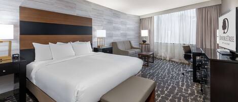 A hotel room with a large bed, a desk, a chair, a bench, and a view of the outdoors.