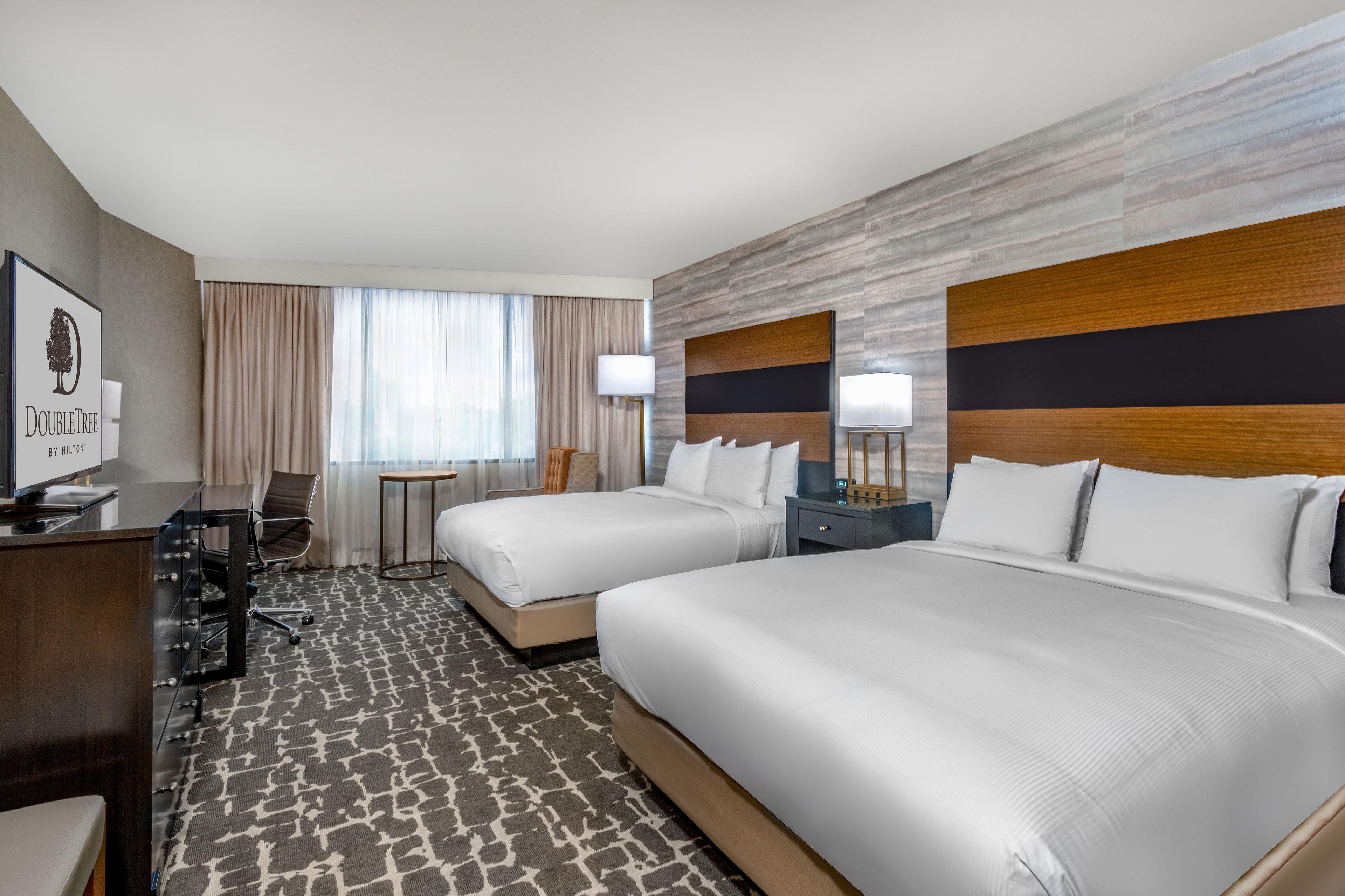 A hotel room with two beds, a desk, a chair, a TV, and a window with curtains.