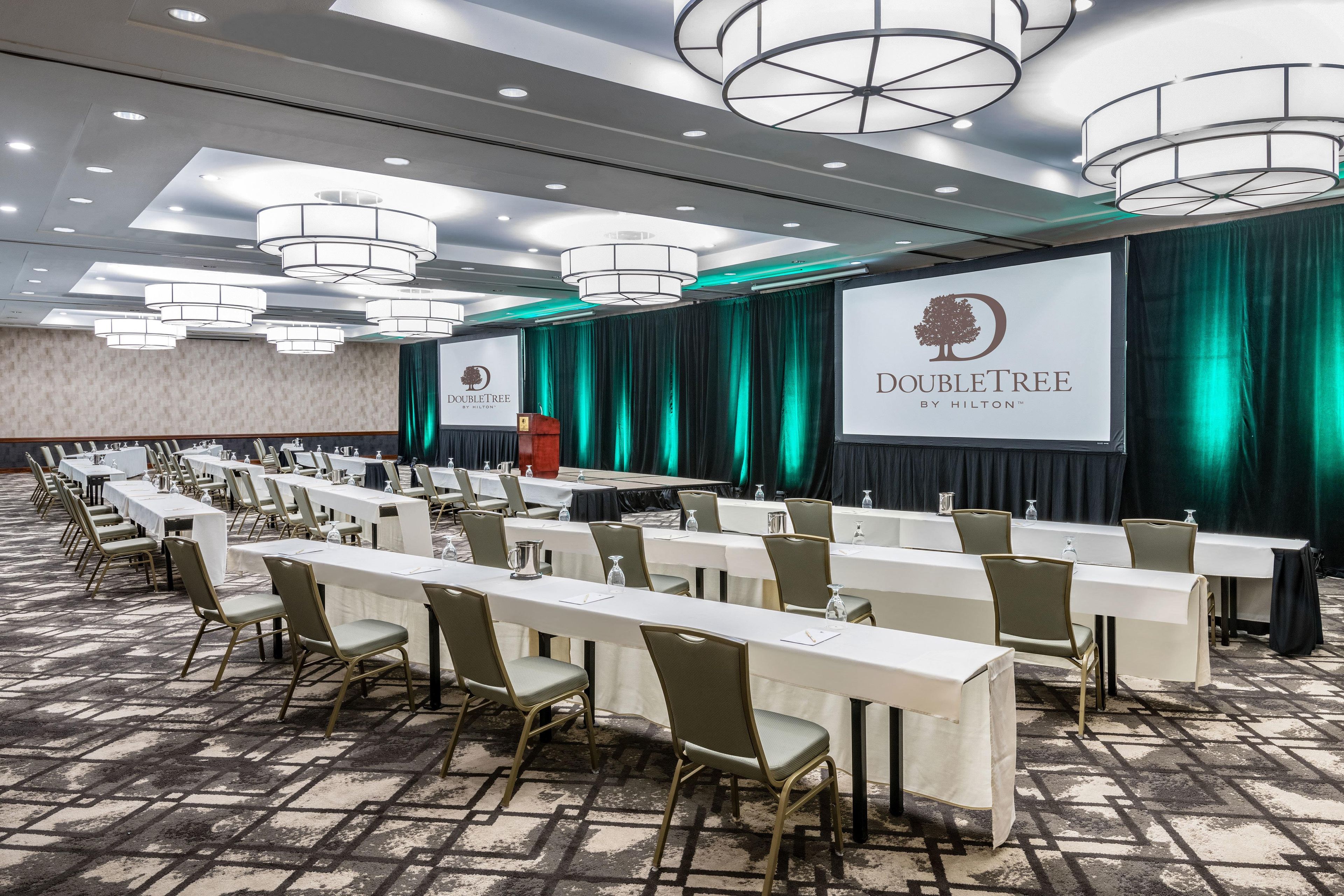 Un salón de conferencias con mesas largas y sillas, un atril y una pantalla grande que muestra el logotipo de DoubleTree.