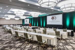 Un salón de conferencias con mesas largas y sillas, un atril y una pantalla grande que muestra el logotipo de DoubleTree.