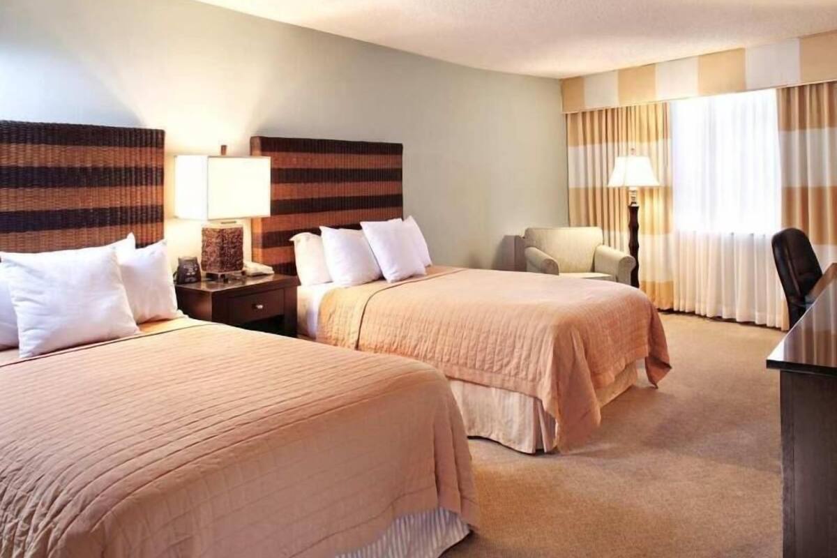 A hotel room with two beds, a desk, a chair, and a lamp.