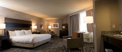 Habitación de hotel con una cama grande, un escritorio, una silla, una lámpara de escritorio, un televisor, un baño con lavamanos y espejo, y piso alfombrado.