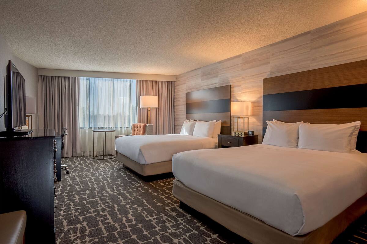 A hotel room with two beds, a desk, a chair, a lamp, and a view of the outdoors.