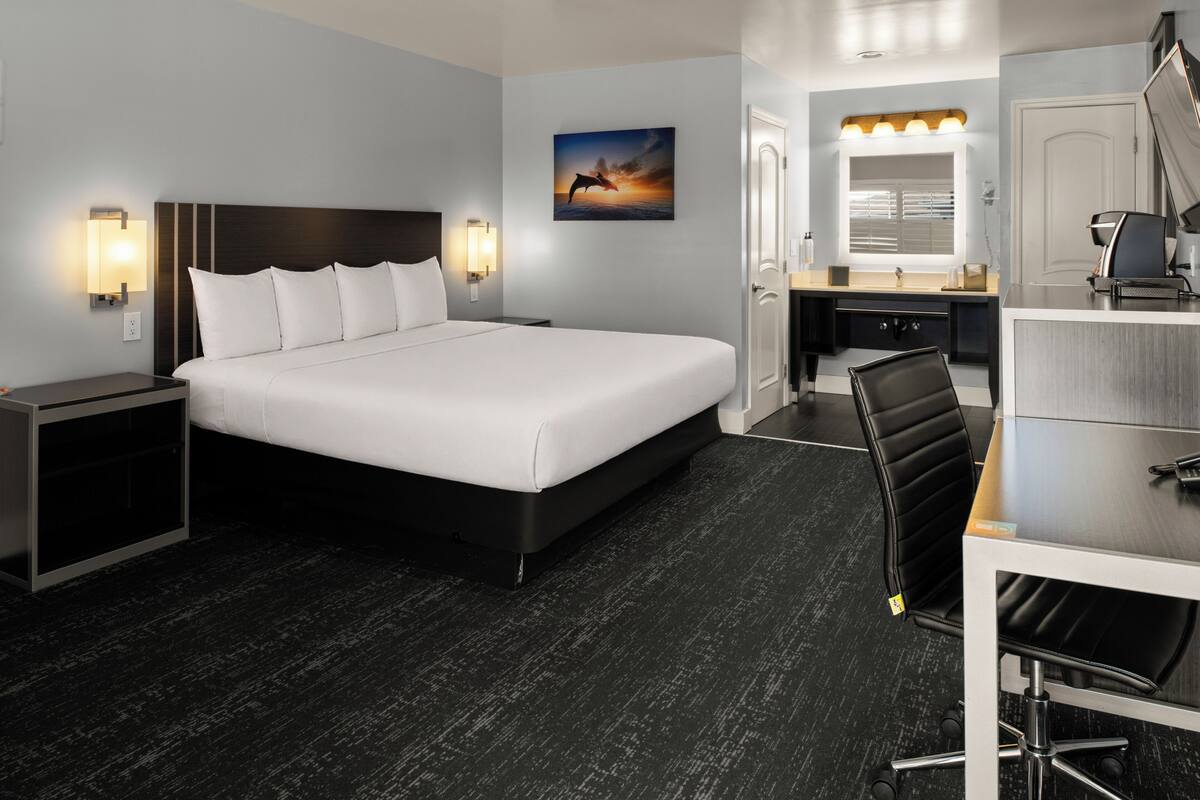 A hotel room with a large bed, a desk, a chair, a desk lamp, a nightstand, a wall-mounted light, and a painting of a sunset.