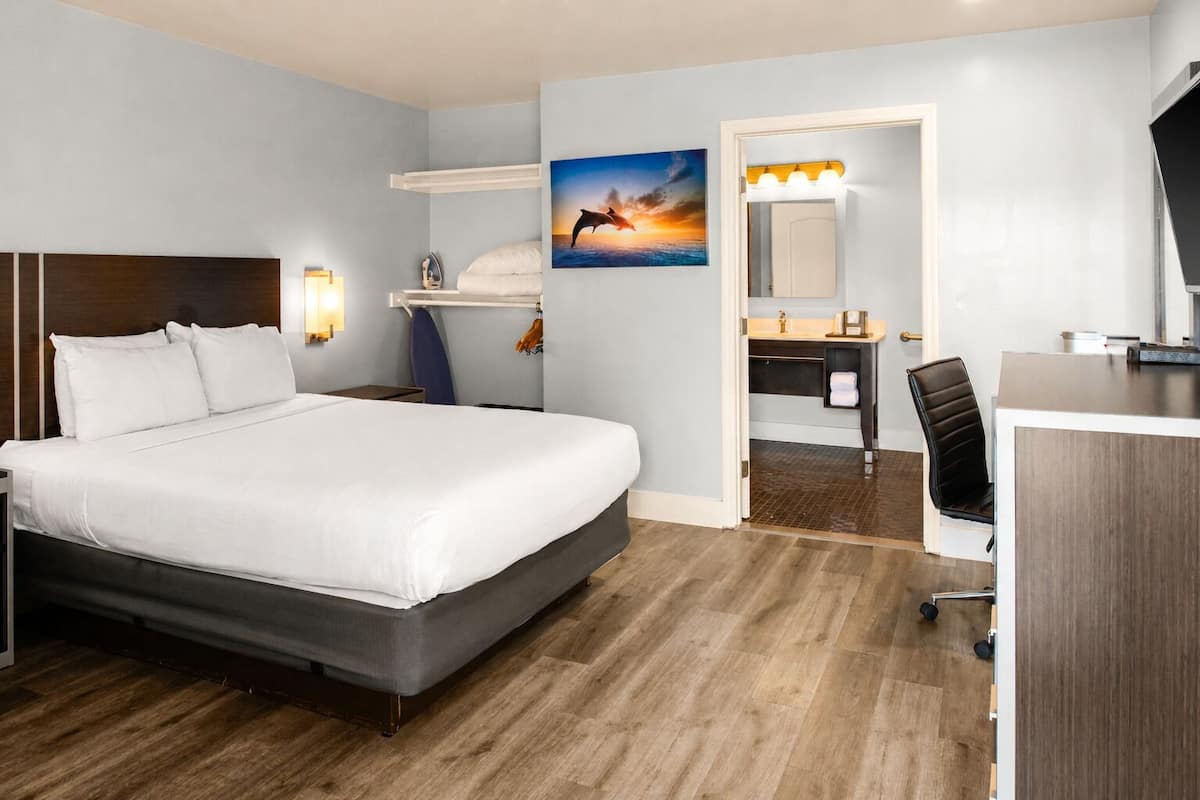A hotel room with a large bed, a desk, a chair, a bathroom with a mirror, and a painting on the wall.