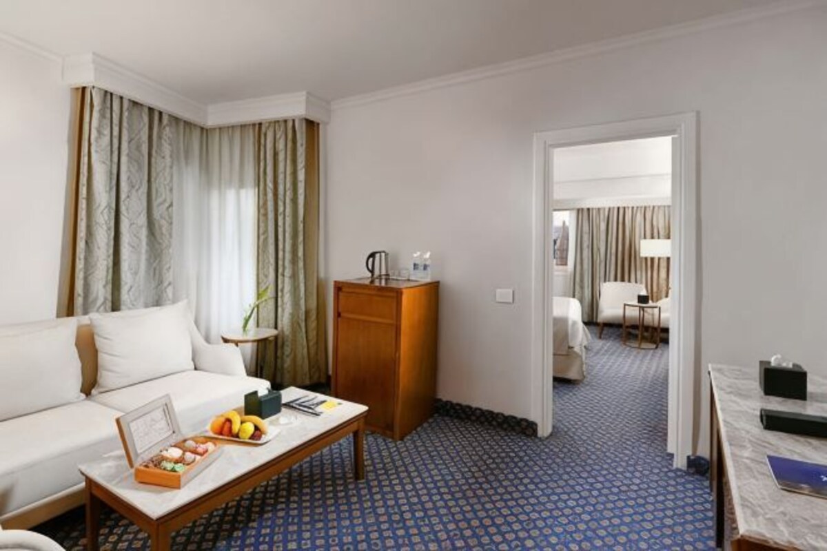 A hotel room with a sofa, a coffee table with fruits, a desk, and a bed in the background.