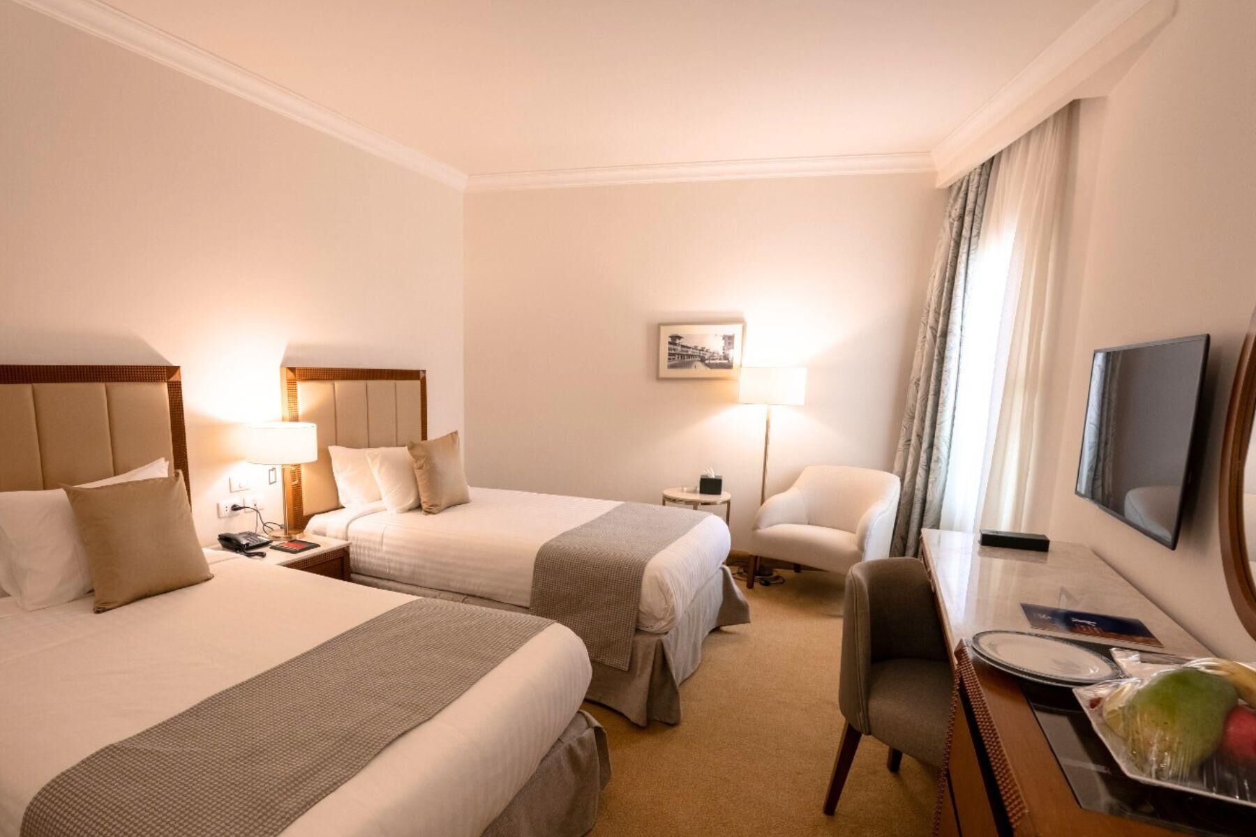 A hotel room with two beds, a desk, a chair, a TV, and a small table with a remote control and a book.