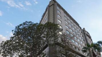 Un hotel de varios pisos con un diseño moderno, que presenta una entrada prominente en esquina y un área ajardinada con árboles.